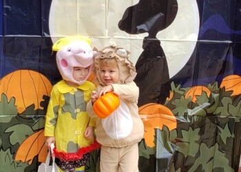 Two children in costumes stand in front of a Halloween backdrop with a full moon and pumpkin patch. One child is dressed as a pig and the other as a lion, holding an orange pumpkin. Several pumpkins are on the ground nearby.