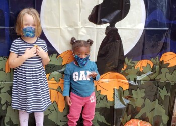 Two young children wearing face masks stand in front of a Halloween backdrop featuring a large moon and cartoon dog silhouette. Several pumpkins and gourds are arranged on the ground beside them.