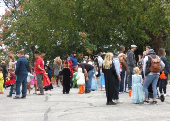 A group of adults and children, many in costumes, gather outdoors on a paved area under large trees during a fall event or Halloween celebration. Some children hold small orange buckets.