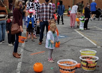 Children in Halloween costumes stand in line at an outdoor event, playing a tossing game with baskets and orange plastic pumpkins. Adults nearby supervise, some wearing masks. Brick building in the background.