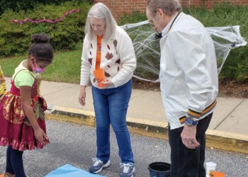 A young girl in costume plays a ball-toss game with two older adults outdoors. The adults hold orange cups and watch as she aims for cups on the ground. Halloween decorations are visible in the background.