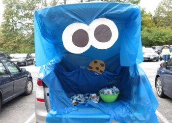 A car trunk decorated as Cookie Monster with large cartoon eyes, a blue tarp for the face, a paper cookie near the mouth, and a bowl of cookies inside, set in a parking lot.
