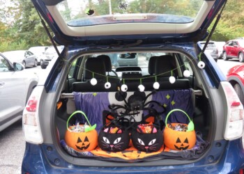 An open car trunk decorated for Halloween with pumpkin and black cat candy buckets, a large spider, a purple spooky cloth, and hanging eyeball decorations. The trunk is filled with candy, parked in a lot.