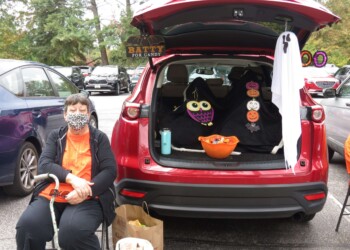 A woman wearing a face mask sits beside a red SUV with its trunk open, decorated for Halloween with owl and pumpkin decorations, a ghost, and a sign reading Batty for Candy. Bowls of candy sit in the trunk and on the ground.
