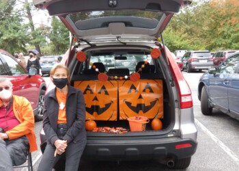 Two people sit by an open SUV trunk decorated with large jack-o’-lantern faces, pumpkins, and Halloween-themed items in a parking lot. Both are wearing jackets and face masks.