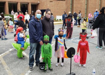 Children and adults, many in Halloween costumes and face masks, gather outdoors in a parking lot by a brick building for a festive event. Some children hold treat buckets, with parents standing nearby.