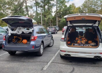 Two SUVs with open trunks decorated for Halloween are parked side by side in a lot. The trunks display pumpkins, fake cobwebs, skeletons, and Halloween signs, with trees and other cars in the background.
