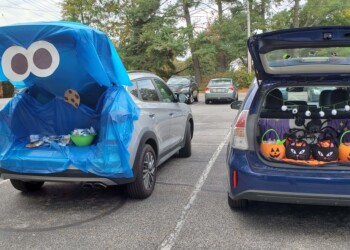 Two cars in a parking lot with trunks open for a Halloween event. One trunk is decorated as Cookie Monster with blue fabric and big eyes, the other has pumpkin buckets and purple cobweb decorations inside.