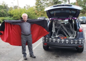 A man dressed as a vampire stands next to an SUV with its trunk open, decorated with cobwebs, skull garland, and Halloween candy inside. The scene takes place in a parking lot with trees in the background.