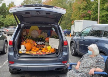 A woman in a mask sits next to an SUV with its trunk open, decorated for Halloween with pumpkins, a Snoopy plush, and Peanuts characters. A sign reads Welcome Great Pumpkin. Other cars are visible in the parking lot.