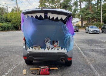 A car decorated for Halloween with its trunk open, draped in blue and purple fabric and lined with large white teeth to look like a monster’s mouth. Nearby, tools and a red tool bag sit on the ground in the parking lot.