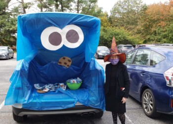 A car decorated with blue fabric to look like Cookie Monster, featuring large eyes and a cookie on the front. A person in a witch costume and mask stands nearby, with another decorated car next to them.