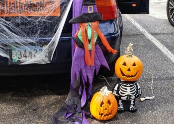A car decorated for Halloween with a spider, web, “Happy Halloween” sign, and costumed pumpkins—one as a witch, one as a skeleton, and a carved jack-o’-lantern. A woman stands by the open car door.