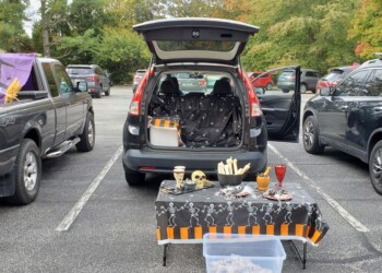 A Halloween-themed trunk-or-treat setup in a parking lot features a decorated car trunk with skeletons, skulls, bones, a table with spooky decorations, and a plastic tub in front. Other decorated cars are parked nearby.