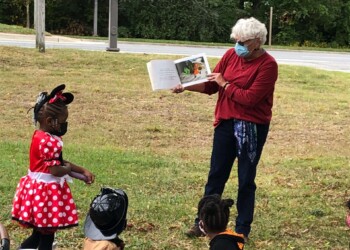 A woman wearing a mask reads a picture book to children dressed in costumes, including Minnie Mouse, a firefighter, and a witch, outside on a grassy area near a road.