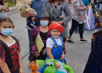 A group of children and adults in Halloween costumes, some wearing masks, stand outdoors on a street. The kids hold colorful trick-or-treat buckets, and trees with green leaves are in the background.
