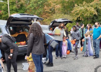 A group of adults and children, some in costumes, line up at open car trunks decorated for Halloween in a parking lot. People hold treat buckets and wear face masks. Trees with autumn leaves are visible in the background.