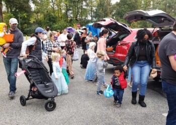 Children and adults in costumes gather in a parking lot for a Halloween trunk-or-treat event. Some kids carry buckets for candy, and several adults hold children or push strollers near open car trunks.