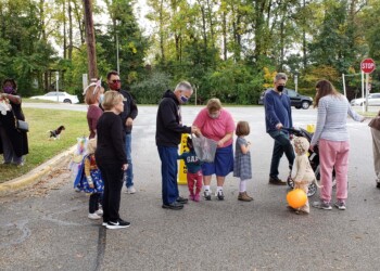 A group of adults and children dressed in costumes gather outdoors near trees, some holding treat bags and pumpkins, participating in a Halloween event on a paved area by a street with stop signs.
