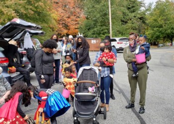 A group of adults and children, some in costumes, gather outdoors near parked cars. One woman holds a baby, others stand or crouch by strollers. Autumn trees and a dumpster are in the background.