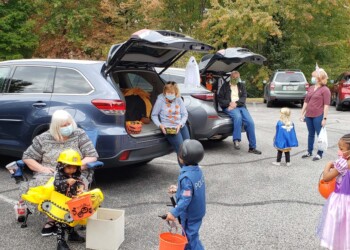 Children in costumes gather around cars with open trunks for trick-or-treating in a parking lot. Adults, some wearing masks, hand out candy while fall trees are visible in the background.