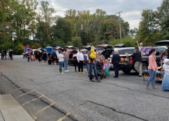 People and children in costumes gather around decorated car trunks in a parking lot for a trunk-or-treat event. Trees and power lines are visible in the background.
