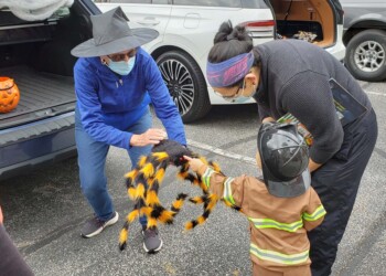 A child in a firefighter costume receives a large, plush spider from an adult in a parking lot. Two adults, one wearing a witch hat, are participating. Halloween decorations and cars are visible in the background.