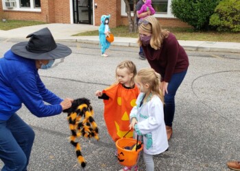 Two young children in Halloween costumes hold pumpkin buckets as a woman in a witch hat shows them a large furry spider. Adults and other children, some in costumes, are nearby on a street outside a brick building.