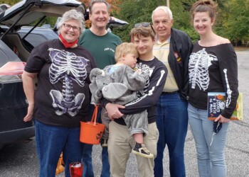 Six people pose outdoors at a Halloween event. Two women wear matching skeleton shirts, a man holds a child in an elephant costume, and two others stand smiling. Cars and autumn trees are in the background. A decorated table is in front.
