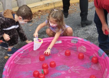 Two young children in masks play by a pink inflatable kiddie pool filled with water and red floating objects. One child uses a net while the other looks on. An adult stands nearby.
