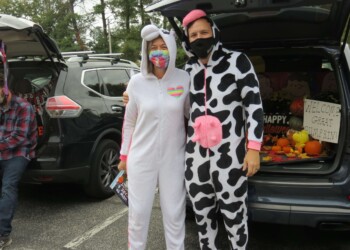 Two people in animal onesies, one as a unicorn and the other as a cow, stand together in a parking lot decorated for Halloween. They both wear face masks, and festive decorations and signs are visible in open car trunks behind them.