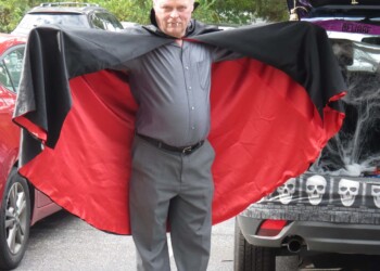 A man dressed as a vampire stands with a black and red cape spread open. He has fangs in his mouth and is standing by a decorated car with skulls and spider webs in a parking lot.