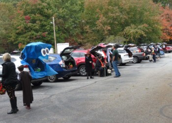 People participate in a trunk-or-treat event in a parking lot, with decorated car trunks and children in costumes collecting candy. Trees with autumn foliage are visible in the background.