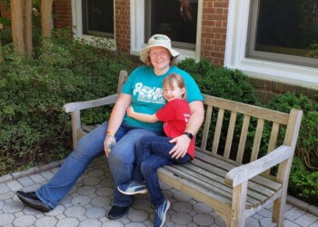 An adult and a young child sit together on a wooden bench outside, smiling. The adult wears a hat and teal shirt; the child wears a red shirt. Behind them are green bushes, brick walls, and large windows.