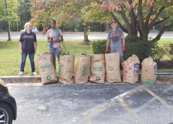Three people stand outdoors behind a row of large paper lawn bags filled with yard debris. They are smiling, wearing casual clothes and gloves, with trees and a parking lot in the background.