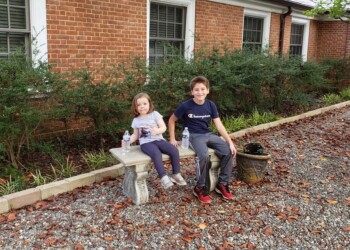 Two children sit on a stone bench in a garden with gravel and fallen leaves, in front of a red brick house with bushes and windows. One child holds a water bottle; both are smiling at the camera.
