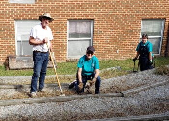 Three people are working outdoors in a garden area beside a brick building. One person stands holding a rake, while two others kneel and squat, wearing gloves and using gardening tools.