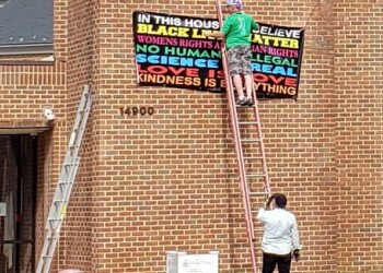 A person stands on a ladder hanging a colorful sign on a brick building, while another person watches from below. The sign lists social justice messages, including “Black Lives Matter” and “Love is Love.”.