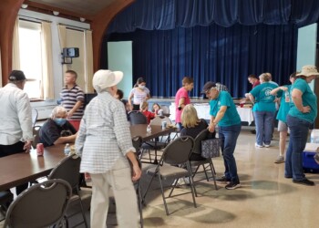 A group of people gather in a community hall with tables and chairs. Some are seated, while others stand or walk around. The atmosphere appears casual and social, with a stage and blue curtain in the background.