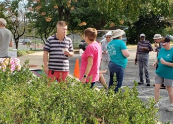 A group of people gather outdoors near green bushes, some standing and chatting while others enjoy ice cream. Trees and parked cars are visible in the background on a sunny day.
