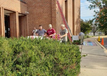 Three people stand near a table with a colorful cloth outside a brick building, partially obscured by bushes. A sidewalk, ladder, and parked car are visible, along with autumn trees and a bright sky.