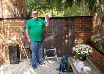 A man in a green shirt stands smiling next to a step stool in an outdoor memorial area with engraved plaques on a brick and stone wall, holding a white cloth. A bench with a backpack and flowers is nearby.
