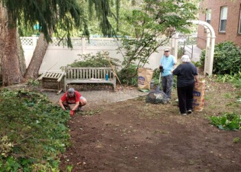 Three people work in a garden: one kneels pulling weeds, while two others stand near large paper yard waste bags. Nearby are trees, shrubs, a wooden bench, and a brick building in the background.