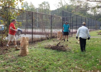 Three people clean up a grassy area next to a tennis court, collecting sticks and yard debris into brown paper bags. Trees and a chain-link fence are in the background.